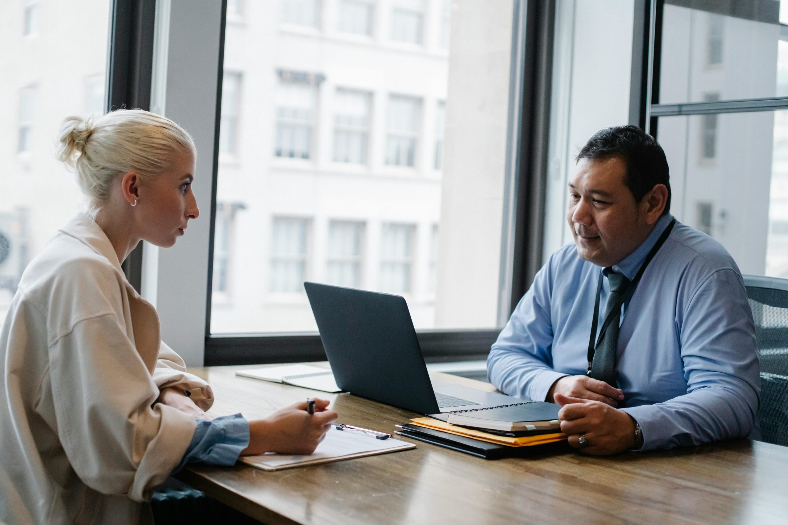Man in a blue shirt and tie talks with a colleague taking notes at a desk with a laptop.